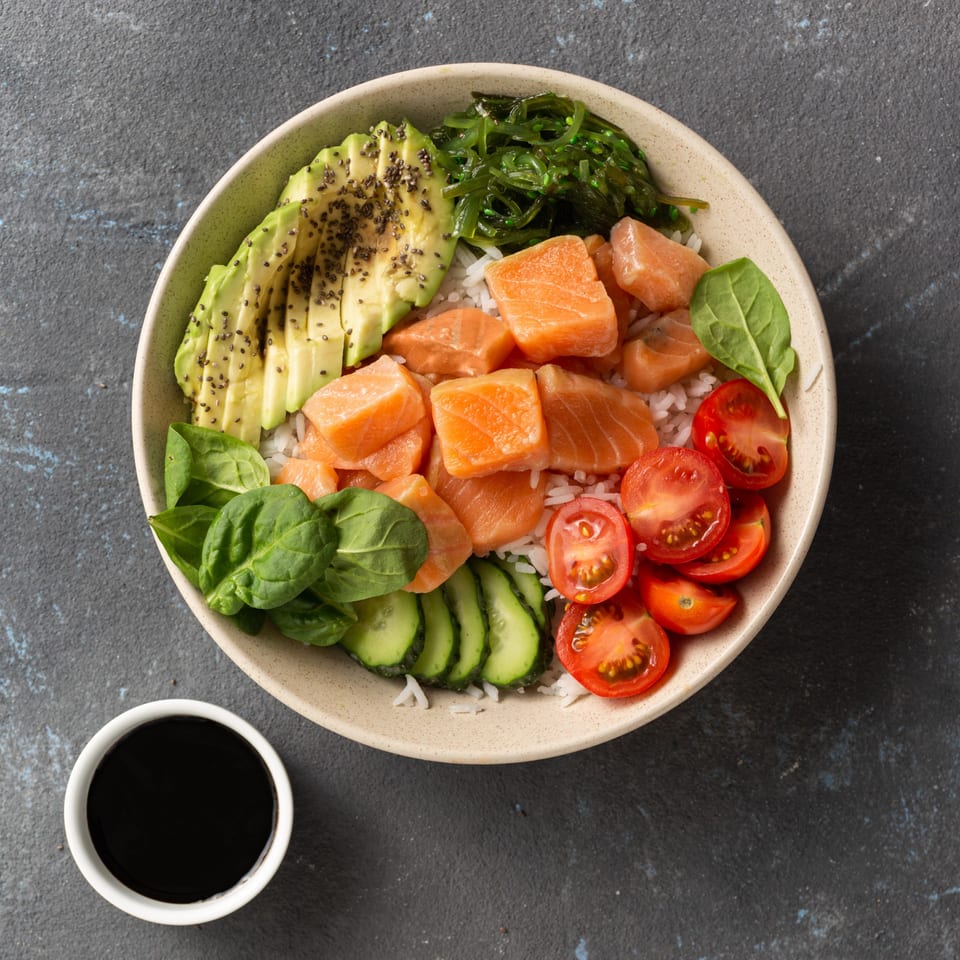 A colorful poke bowl with salmon, avocado, seaweed, tomatoes, spinach, and cucumber, with a side of soy sauce.